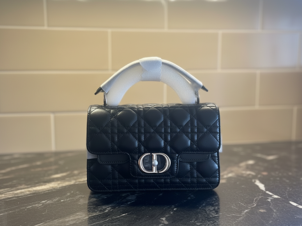 A black quilted leather handbag with flap top closure, a leather top handle, and long silver metal chain strap, featuring a silver-tone Dior logo with pearl accent clasp.  The bag is resting on a dark granite countertop with a beige tiled wall in the background.