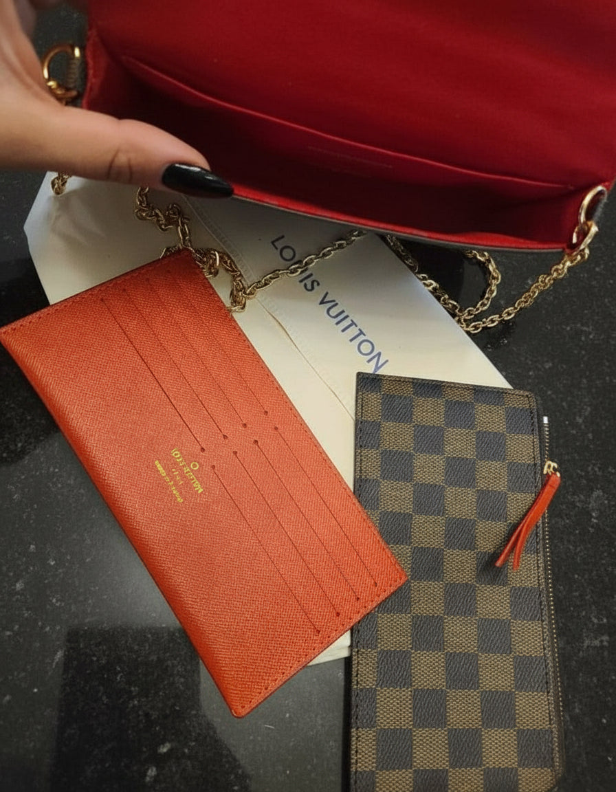 Red handbag with a gold chain, orange wallet, and checkered pouch on a dark surface.