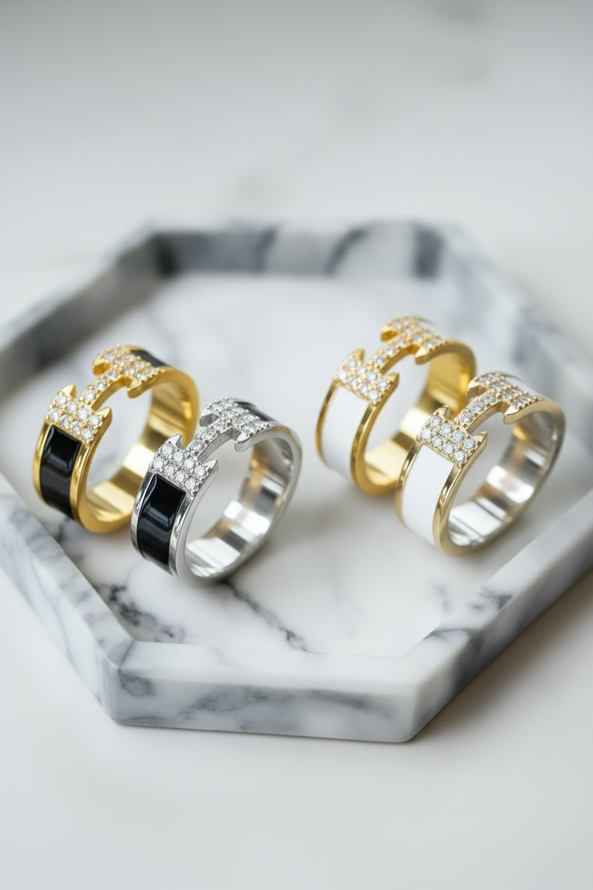 Four stylish adjustable thick band rings. Two are Gold and two are silver, each has the distinctive letter H logo with crystals in the center.  Two of the rings have white bands and the other two have black bands. 