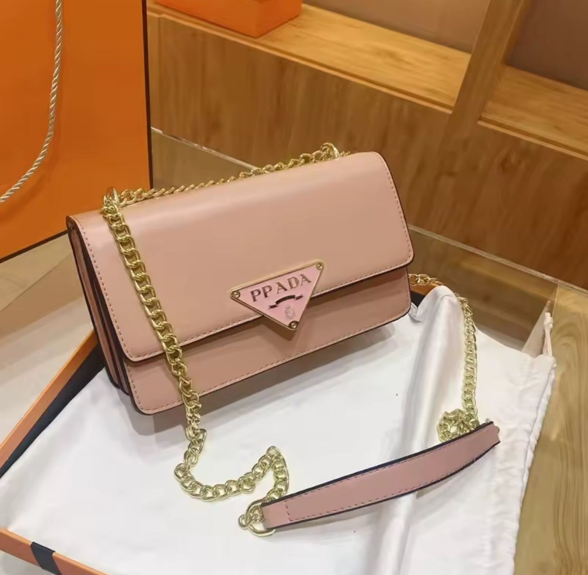 Pink handbag with gold chain on a white surface, with an orange box in the background.